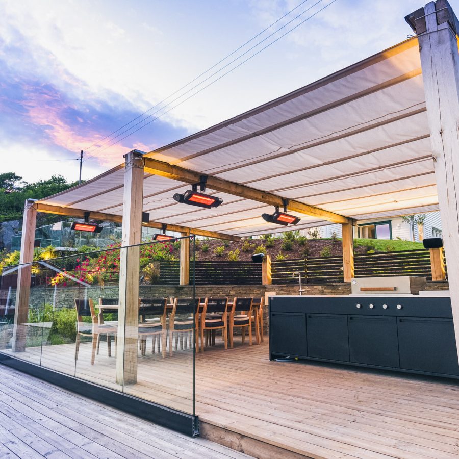 Stor terrasse i vakkert naturlandskap med pergola, stor sittegruppe, utekjøkken og flere varmelamper i taket.